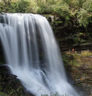 A wide waterfall cascades over a rocky ledge surrounded by lush greenery, with a wooden walkway and people admiring the stunning waterfall near the base.