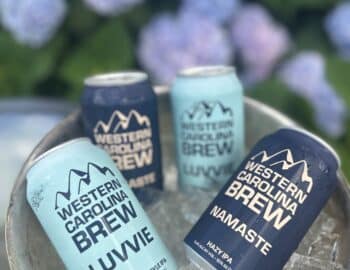 Four cans of Western Carolina Brew in blue and navy colors labeled “Luvvie” and “Namaste” sit in a metal bucket filled with ice, with hydrangeas in the background.